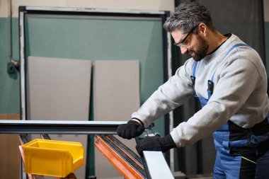 Orta yaşlı bir işçi atölyede pencere profili yapıyor. Koruyucu eldiven takıyor.
