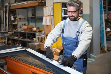 Atölyede elinde matkapla gülümseyen adam, güvenlik gözlüğü takıyor.