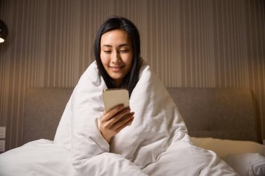 Gülümseyen bir kadın evde eğlenmekten zevk alıyor battaniyeye sarılmış bir şekilde yatakta oturuyor ve sosyal ağlarda akıllı telefonlarla sohbet ediyor ya da internetten mesaj atıyor, oyun oynuyor.