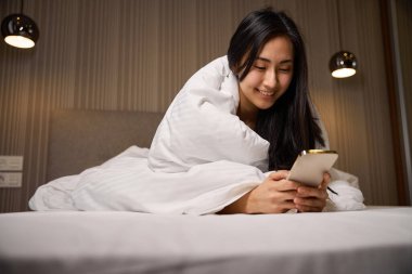 Battaniyeye sarılı gülümseyen kadın rahat bir yatağa uzanıp akıllı telefondan özel uygulama kullanarak ürünlerin teslimatını emrediyor.