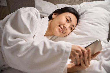 Beyaz yumuşak bornozla yatakta uzanmış, arkadaşlarıyla ya da sevgilileriyle akıllı telefondan sohbet eden, Asyalı kökenli gülümseyen bir kadın hoş yorumlar okuyor.