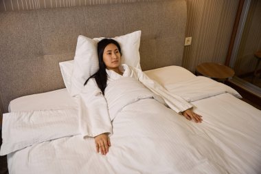 Asyalı etnik kökenli çok sinirli bir kadın beyaz bir bornozla yatağa uzanmış, yorganla örtülmüş, ilgisiz, işini kaybetmiş.