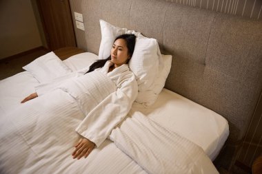 Üzgün Asyalı kadın, beyaz yumuşak bornozla çift kişilik yatakta yatıp bir noktaya bakıyor, ya ayrılık ya da boşanma acısı çekiyor, yorgan ve hüzün içinde.