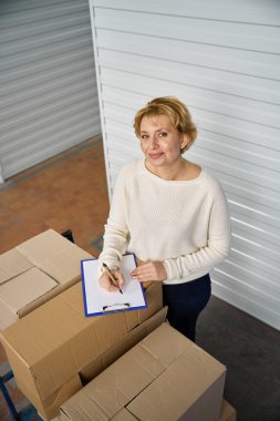 Elinde pano olan bir kadın, depoda kutuların yanında duruyor, kameraya bakıyor ve yazıyor.