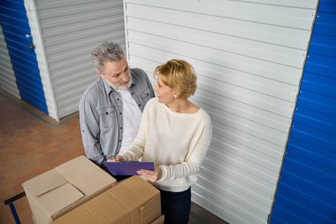 Elinde dosya olan bir kadın, depoda kutuların yanında duruyor. Yazıyor, erkeklere bilgi veriyor.