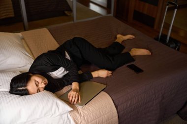 Yorgun bir kadın otel odasında elinde dizüstü bilgisayarla dinleniyor, geç saatlere kadar konuşmasına çalışıyor ve iş elbisesiyle uyuyakalıyor, aşırı çalışıyor.
