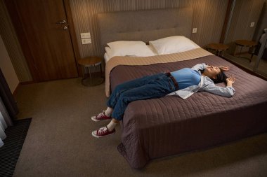 Yan manzara gülümseyen Asyalı kadın otel odasındaki büyük yatakta yatıyor, tatillerde eğleniyor, dinleniyor ve otelin atmosferinin tadını çıkarıyor.