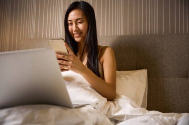 Randevu uygulamasında akıllı telefondan ve laptoptan konuşan gülümseyen Asyalı kadın, arkadaşlarıyla mesajlaşan ya da sosyal ağlarda pijamalarıyla yatağında oturan yorumlar yapan.