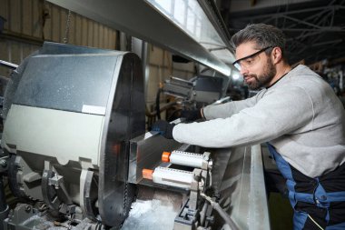 Orta yaşlı erkek, modern ekipman kullanarak, güvenlik gözlüğü ve eldiven takarak alüminyum bir profil çıkarıyor.