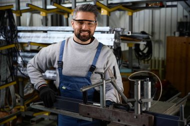 Metal parçası olan modern bir makine üzerinde çalışıyor. Koruyucu eldiven ve gözlük kullanıyor.