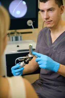 Genç proktolog hastanın önünde otururken nitrile anoskop tutuyor.