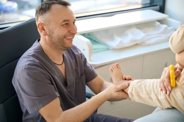 stock image Smiling massotherapist looking at baby seated on mother lap while massaging its calf muscle