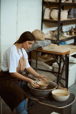 Önlük giymiş esnaf kadın atölye alanındaki ham kilden sofra takımı şekillendiriyor. El işi seramik çömlek üretimi