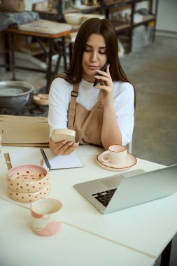 Samimi çömlek atölyesinde oturup sipariş verirken cep telefonu kullanan güzel bir kadın. Küçük işletme kavramı