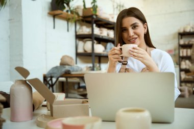 Seramik atölyede kahvenin tadını çıkarırken dizüstü bilgisayarla çalışan gülümseyen genç bayan. Küçük işletme kavramı
