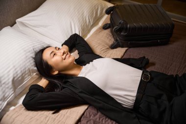 Mutlu Asyalı kadın rahat yatağında uzanıyor ve gülümsüyor, bavulu yakınlarda yatıyor, kadın yönetici uzun iş seyahatinden sonra sonunda evinde dinleniyor.