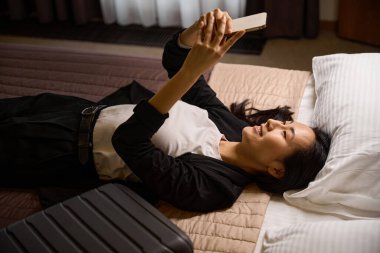 Üst düzey gülümseyen Asyalı kadın akıllı telefonuyla selfie çekiyor otel odasının yatağında uzanıyor, tatillerin tadını çıkarıyor ve sosyal ağlarda fotoğraf paylaşıyor.