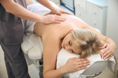 Yetişkin kadın müşteri masadaki yüz üstü yatarken fizyoterapist avuç içi masaj sırasında sırtına bastırıyor.