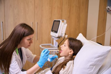 Tedavi gören bir terapist genç bir kızın boğazını inceliyor. Bir kadın steril spatula kullanıyor.