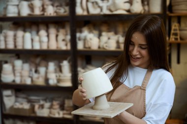 Atölyede elinde vazo tutan önlüklü, gülümseyen genç bir kadın. Çömlek oluşturma süreci