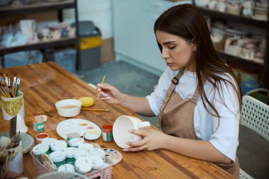 Caucasian young female ceramist painting with brush on clay pottery cup in studio clipart