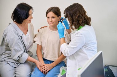 Doktor genç bir kızın kulağını muayene ediyor. Annesi de yanında.