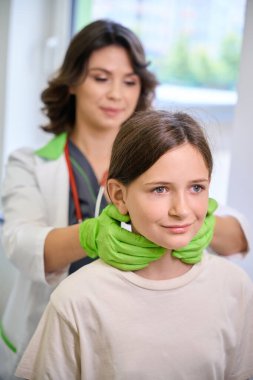 Female pediatrician examines the throat of a teenage girl, the doctor uses protective gloves clipart