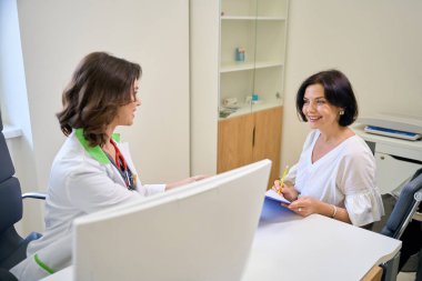 Orta yaşlı bir hasta klinikteki bir randevuda anket imzalar, genç bir doktor kadına tavsiyede bulunur