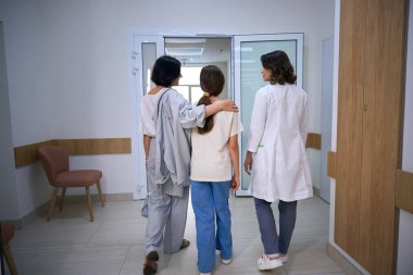 Klinik çalışanı, annesi ile birlikte genç bir hastaya ameliyatta eşlik ediyor. Annesi de kızına sarılıyor.