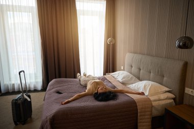 Seyahat kıyafetleri giymiş bir kadın yatağın üzerinde dinleniyor, yanında da seyahat çantası var.