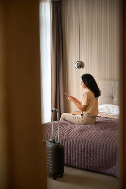 Otel odasında cep telefonu olan orta yaşlı bir kadın, bavulu yakınlarda.