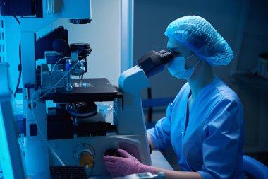 Mavi önlüklü ve koruyucu maskeli bir bilim adamı bir kriyokoruma ünitesinin bilgisayar ekranında çalışıyor.