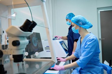 Doktorlar mikroskop ve bilgisayar kurulumu üzerinde birlikte çalışır, örnekleri analiz eder ve laboratuvar işlemlerini yönetirler.