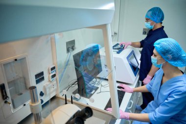 Mavi önlüklü iki laboratuvar teknisyeni embriyo değerlendirme sürecinde büyük bir ekranda görülen analiz sonuçlarını tartışıyor..