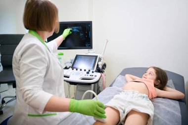 Muayene masasında rahat bir şekilde yatan gülümseyen çocuğa sonuçları açıklamak için ultrason ekranını işaret eden olgun bir doktor..