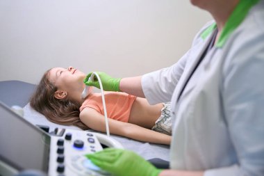 Doktor boynundaki ultrasonu çekerken küçük kız hala masada duruyor. Detaylı görüntülere odaklanıyor.