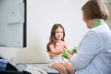 Küçük kız, muayene sırasında doktorla karşılaşıp, çevrede rahat ve rahat bir yaşam sürdüğünü gösterirken ışıl ışıl gülümsüyor.