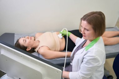 Olgun doktor, yeşil eldiven giyerek, ultrason sondasını nazikçe hastanın karnına yerleştirir, böylece düzgün ve eksiksiz bir muayene sağlar.