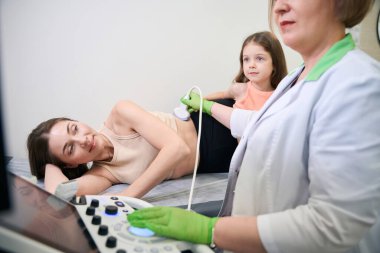 Kadın doktor anne üzerinde karın ultrasonu uygularken, kız da dikkatlice gözlemleyerek ameliyatla ilgilenir.