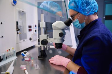 Mavi üniformalı ve koruyucu kıyafetli bir erkek embriyolog örnekleri mikroskopla inceliyor.