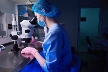 Yumuşak mavi ışığı olan loş bir laboratuvardaki kadın doktorlar, embriyolog örnekleri incelemek için bir mikroskop kullanır.