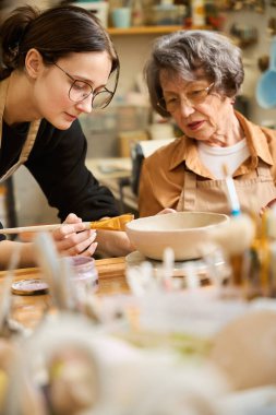 Genç bir kadın yaşlı bir bayana kille nasıl çalışacağını öğretiyor. Stüdyoda ateşlenmemiş işlerle dolu raflar var.