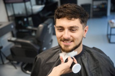 Profesyonel stilist, müşterinin yanında duruyor, büyük fırça tutuyor ve tıraş olduktan sonra pudra sürüyor.