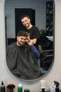 Müşteri aynaya bakıyor yeni saç kesimine, işçinin yanında duruyor, insana bakıyor.