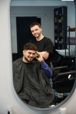 Müşteri aynaya yeni saç kesimine bakıyor, kuaför yanında duruyor, adama bakıyor.