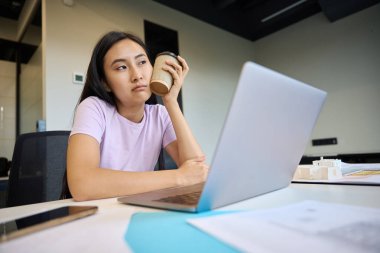 Sad woman is sitting in a chair in front of a laptop, she is tired from a lot of work clipart