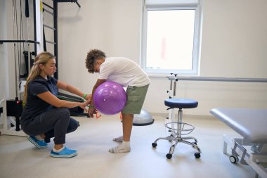 Genç hasta bacak dayanıklılığını ve istikrarını artırmak için terapistin gözetiminde egzersiz topu kullanarak çömelme egzersizi yapıyor.