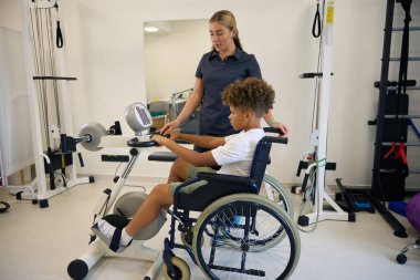 Genç hasta tekerlekli sandalyede üst vücut egzersizleri yaparken terapist de onu denetler ve dayanıklılık ve dayanıklılık üzerine yoğunlaşır.