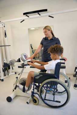 Kadın fizyoterapist, rehabilitasyon merkezinde kol bisikleti kullanırken hastaya destek ve motivasyon sağlayarak egzersiz rutinlerinde yardımcı olur.