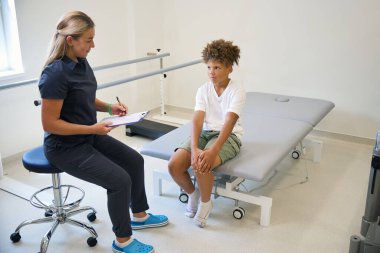 Profesyonel fizyoterapist, genç hastayla fiziksel rehabilitasyon için talimatlar ve teşvik sunan bir egzersiz rutinini tartışıyor